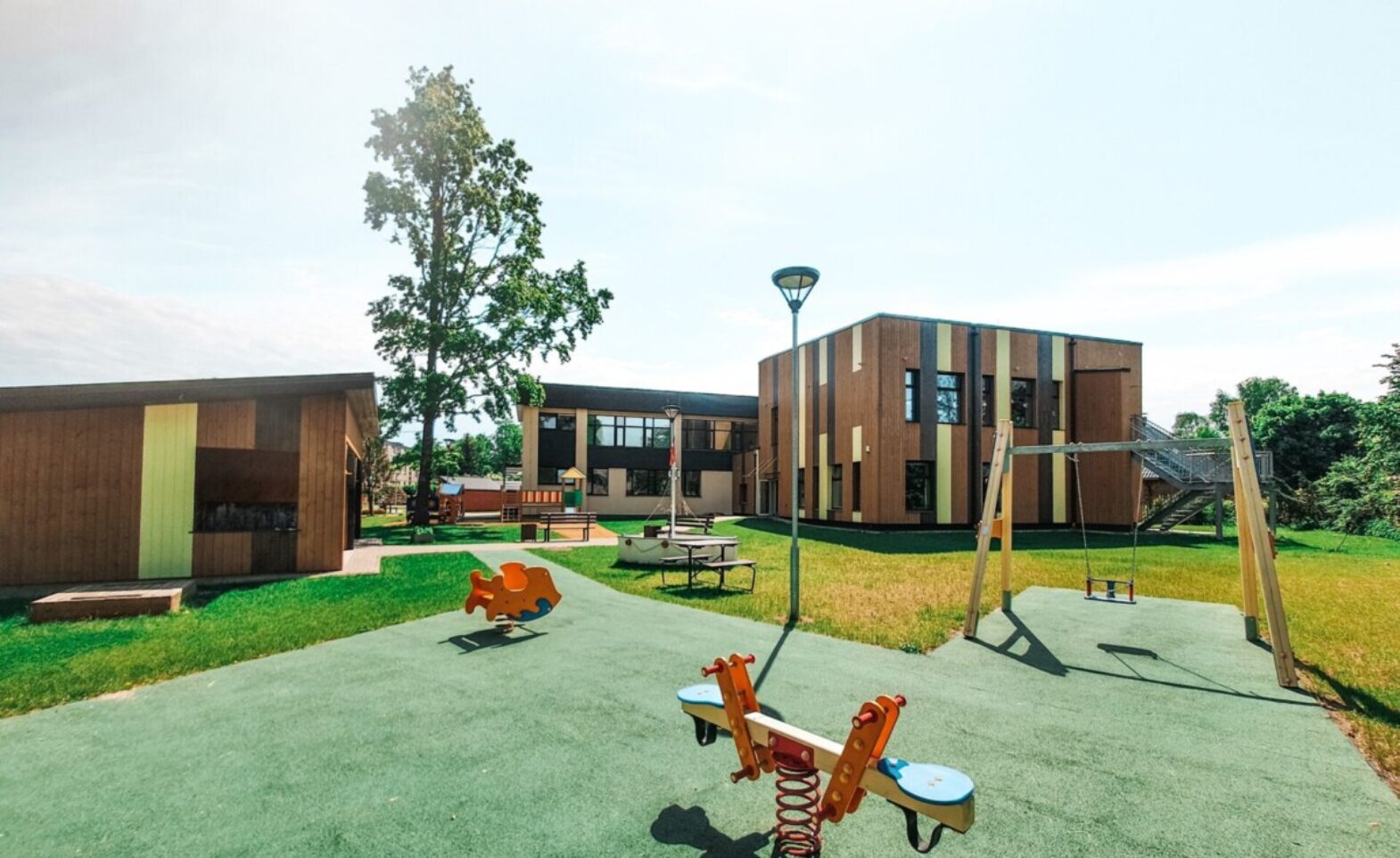 Pirmsskolas spēļu laukums ar šūpolēm, fonā modernas pirmsskolas ēkas.