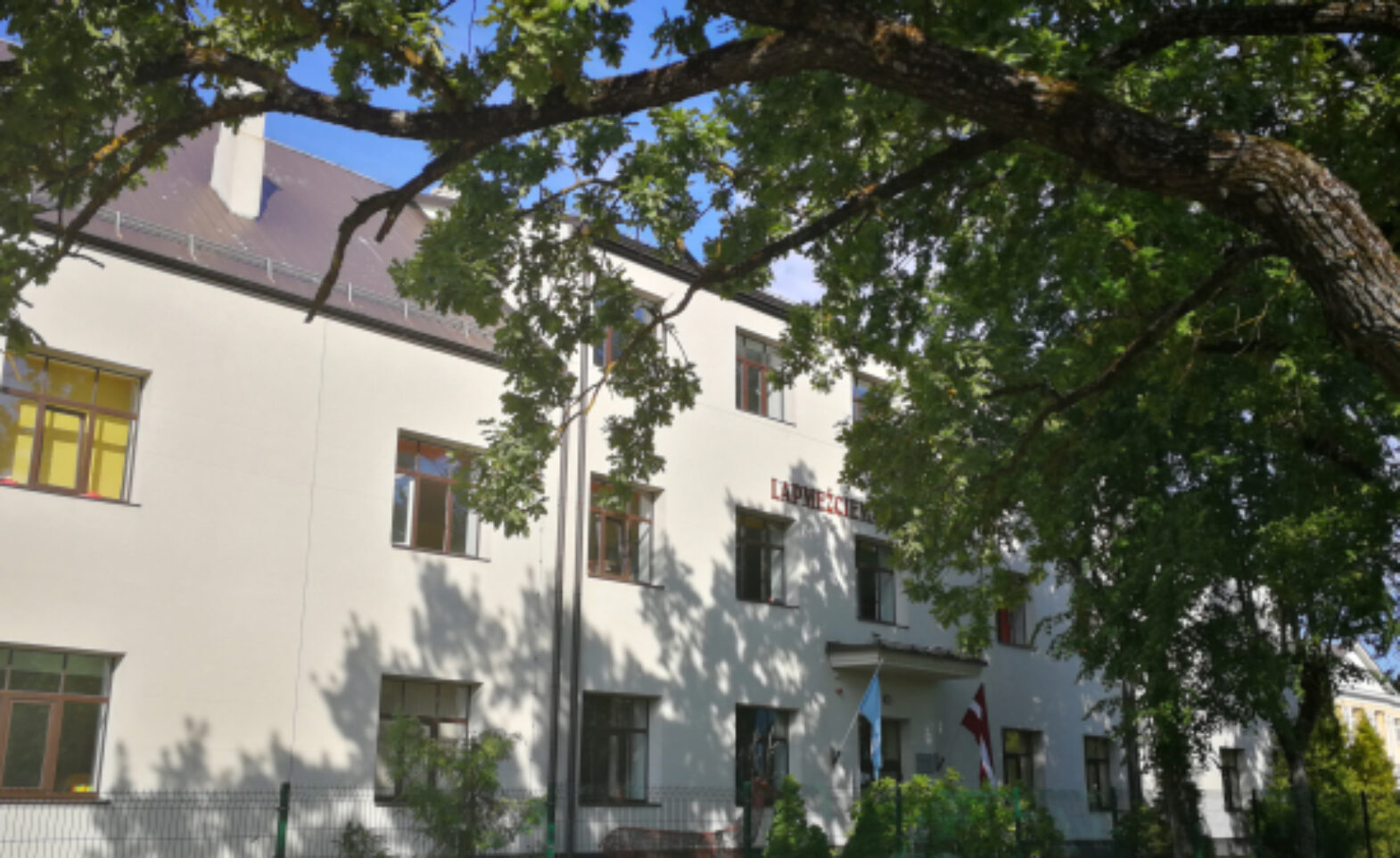 Skolas fasādes foto. Ēku apspīd saule un pie skolas aug ozoli.