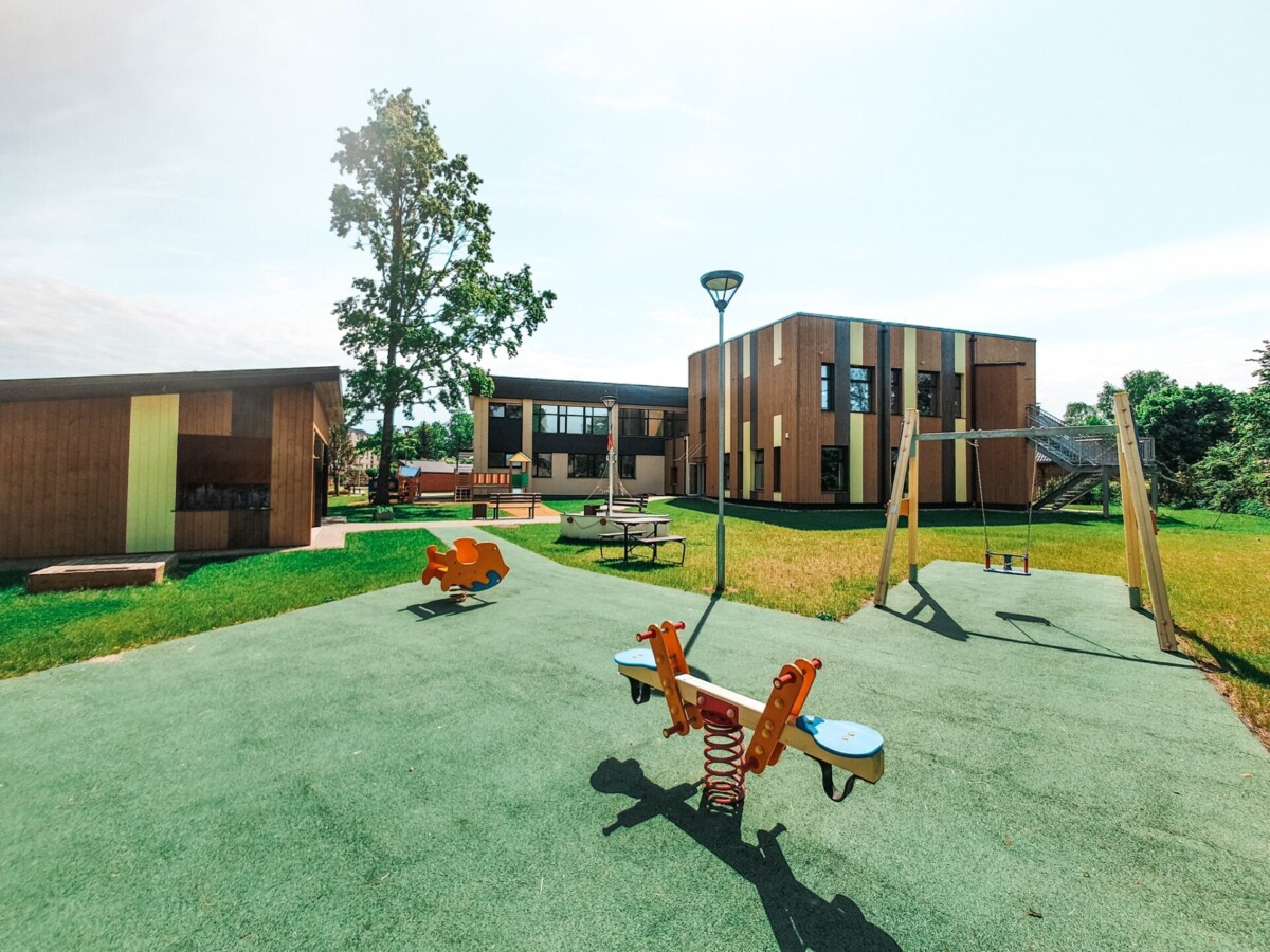 Pirmsskolas spēļu laukums ar šūpolēm, fonā modernas pirmsskolas ēkas.