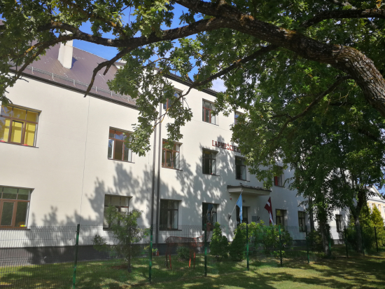 Skolas fasādes foto. Ēku apspīd saule un pie skolas aug ozoli.